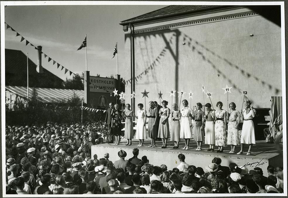 Pent kledte jenter står på en scene med hvert sitt nummerskilt. Folkemengde foran scenen. - Klikk for stort bilde