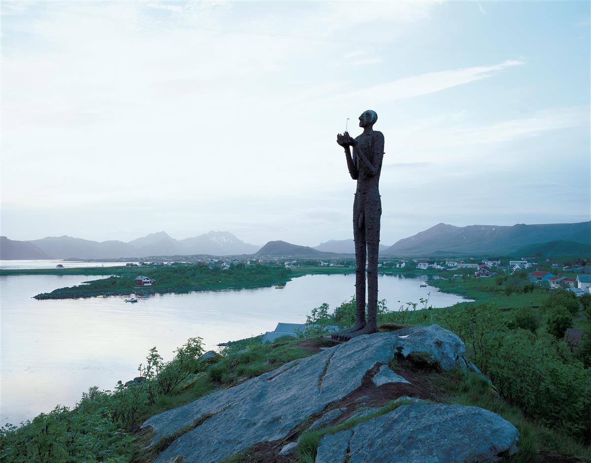 4 meter høy skulptur av en mann som holder en figur i hendene. I bakgrunnen bebyggelse, hav og fjell. - Klikk for stort bilde