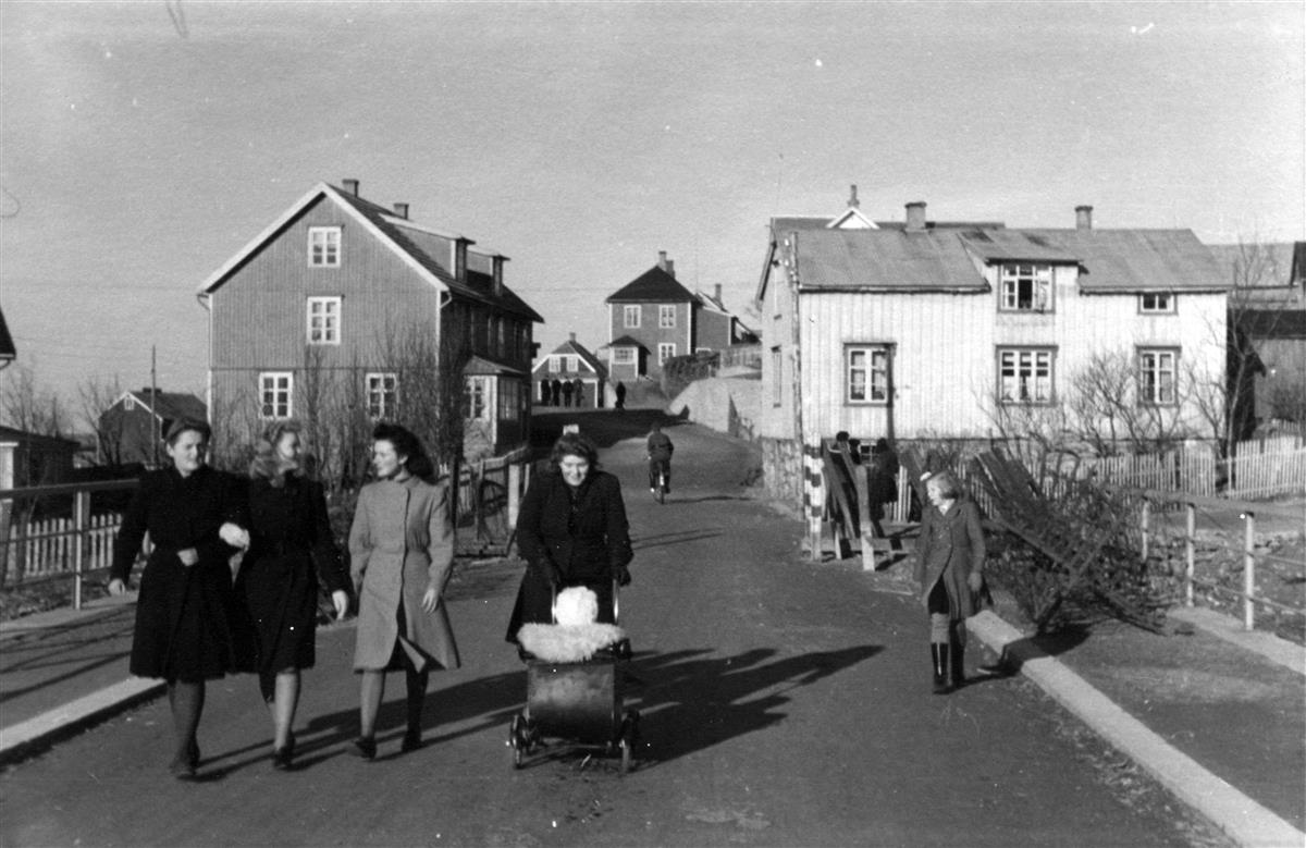 Gatemiljø med trehusbebyggelse. Fire kvinner spaserer med kåpe og småsko over ei bru. Ei barnevogn, ei jente og flere mennesker i bakgrunnen. - Klikk for stort bilde
