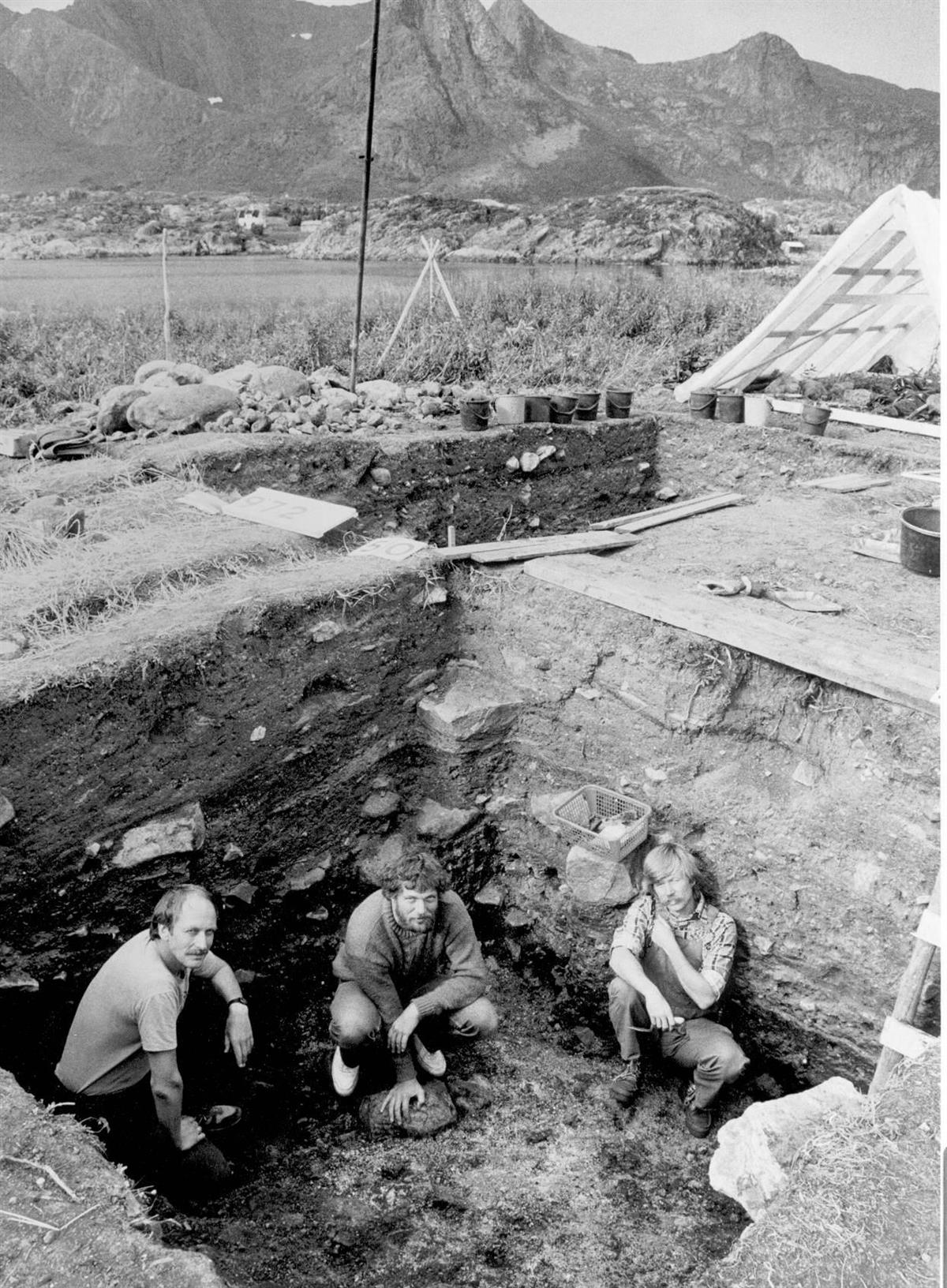 Tre menn i bunnen av en ca to meter dyp utgravingsgrop.  Bøtter, stiger og annet verktøy for utgravingen ses rundt og i gropa. I bakgrunnen fjell og hav. - Klikk for stort bilde
